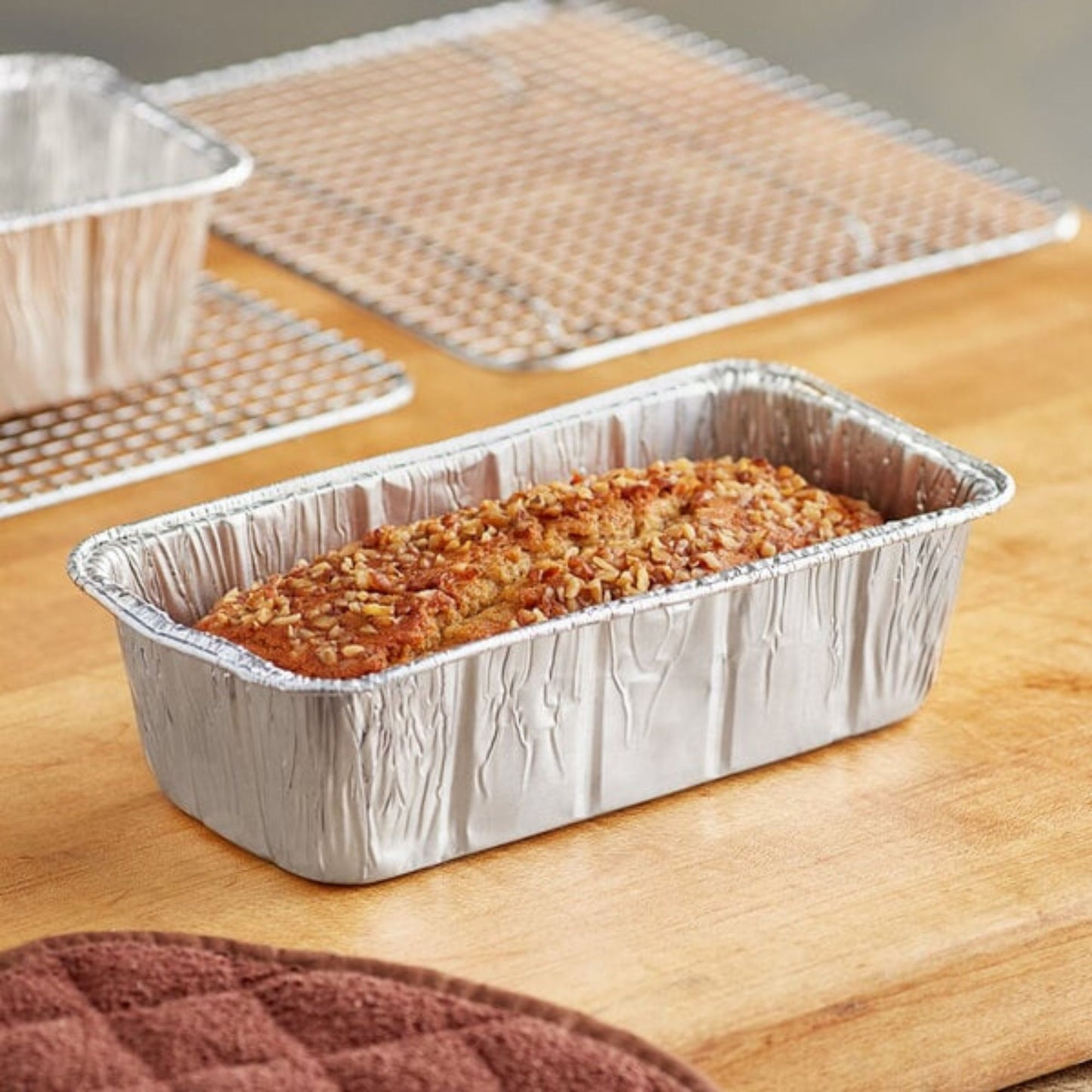 Loaf of bread in a foil pan on a wooden surface with baking trays in the background.