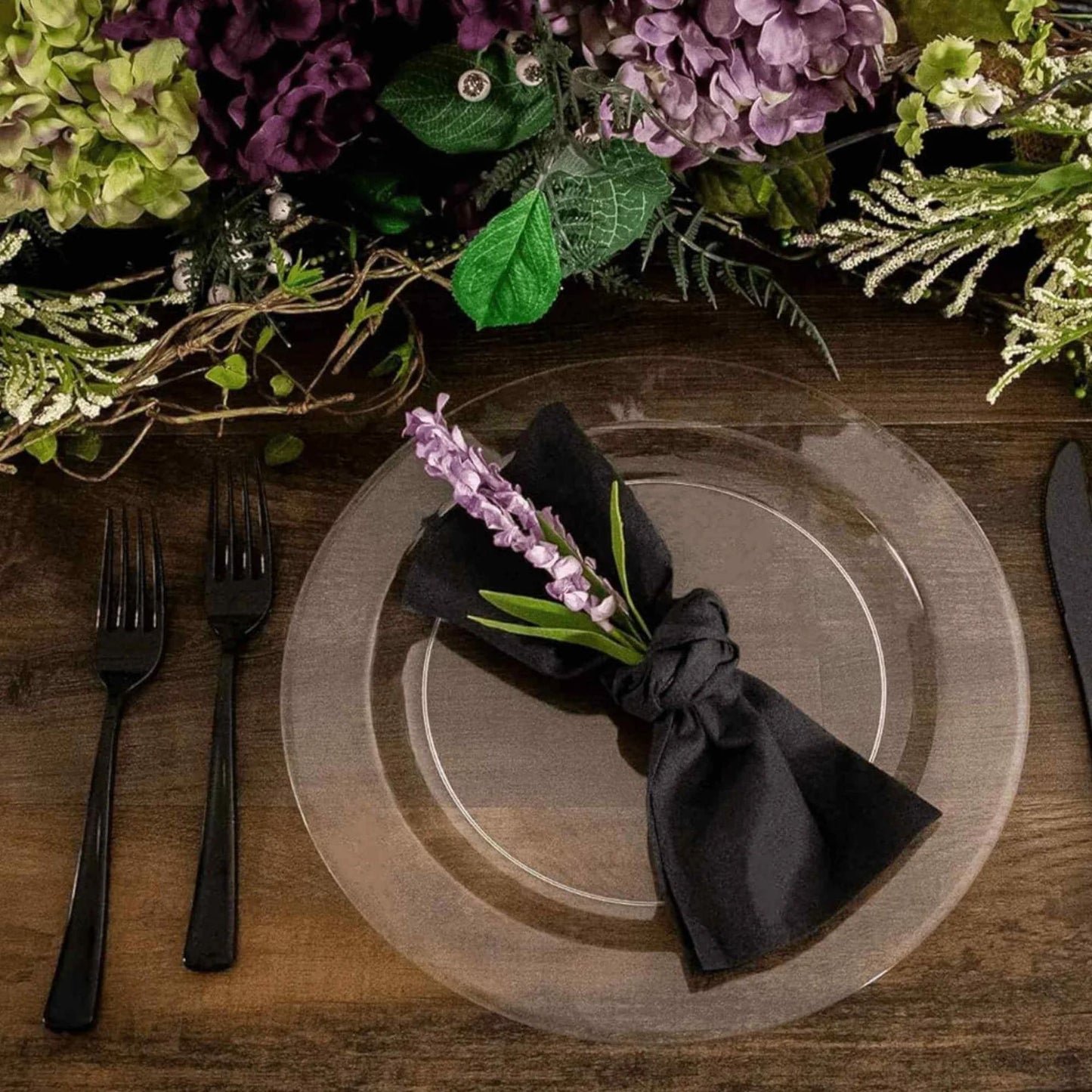 Set table with clear plates, black napkin, and purple flowers on a wooden surface.