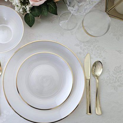 Elegant table setting with white plates, gold rims, silver cutlery, and floral decorations on a textured surface.